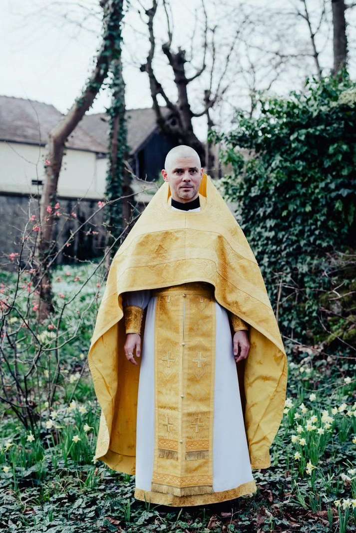 Full shot of an adult male in religious vestments, standing outdoors among greenery with white flowers and a building in the background.