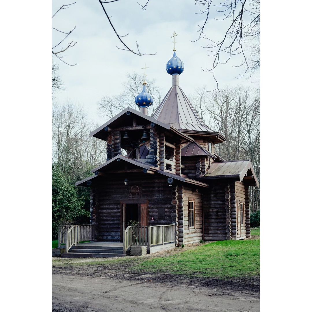 Eastern Orthodox church exterior featuring a log construction style, set against a backdrop of trees and a cloudy sky in a daylight.