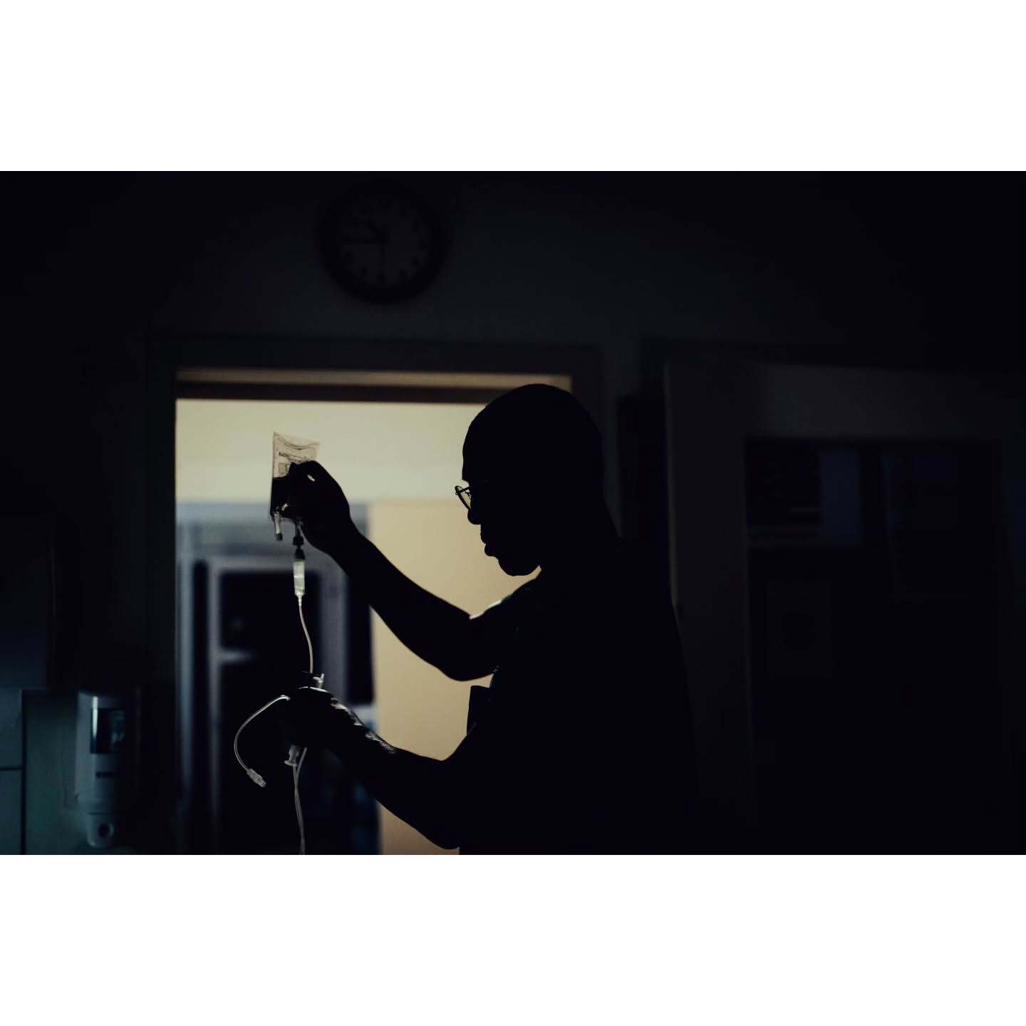 Silhouette of an adult in glasses holding an IV bag, medical equipment, and a clock on the wall inside a dimly lit hospital room.