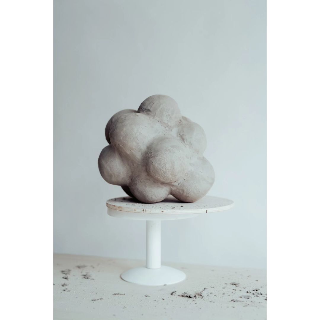 Abstract art featuring a light gray, bulbous sculpture on a white stand against a light gray background, creating a minimalist aesthetic.