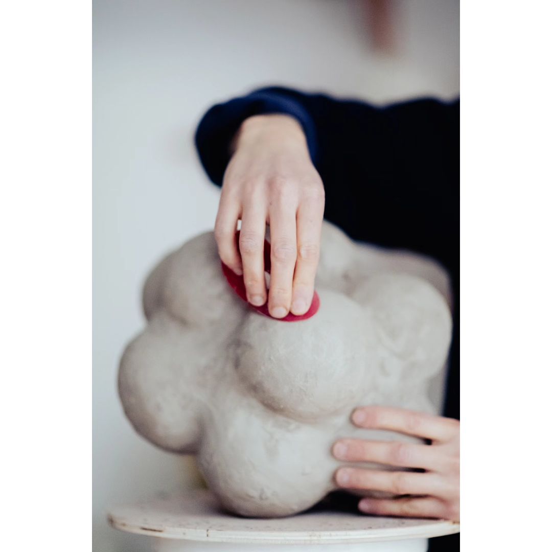 An adult works with clay, shaping a sculpture, while using a red shaping tool in an indoor environment with soft lighting. Focus on hands.