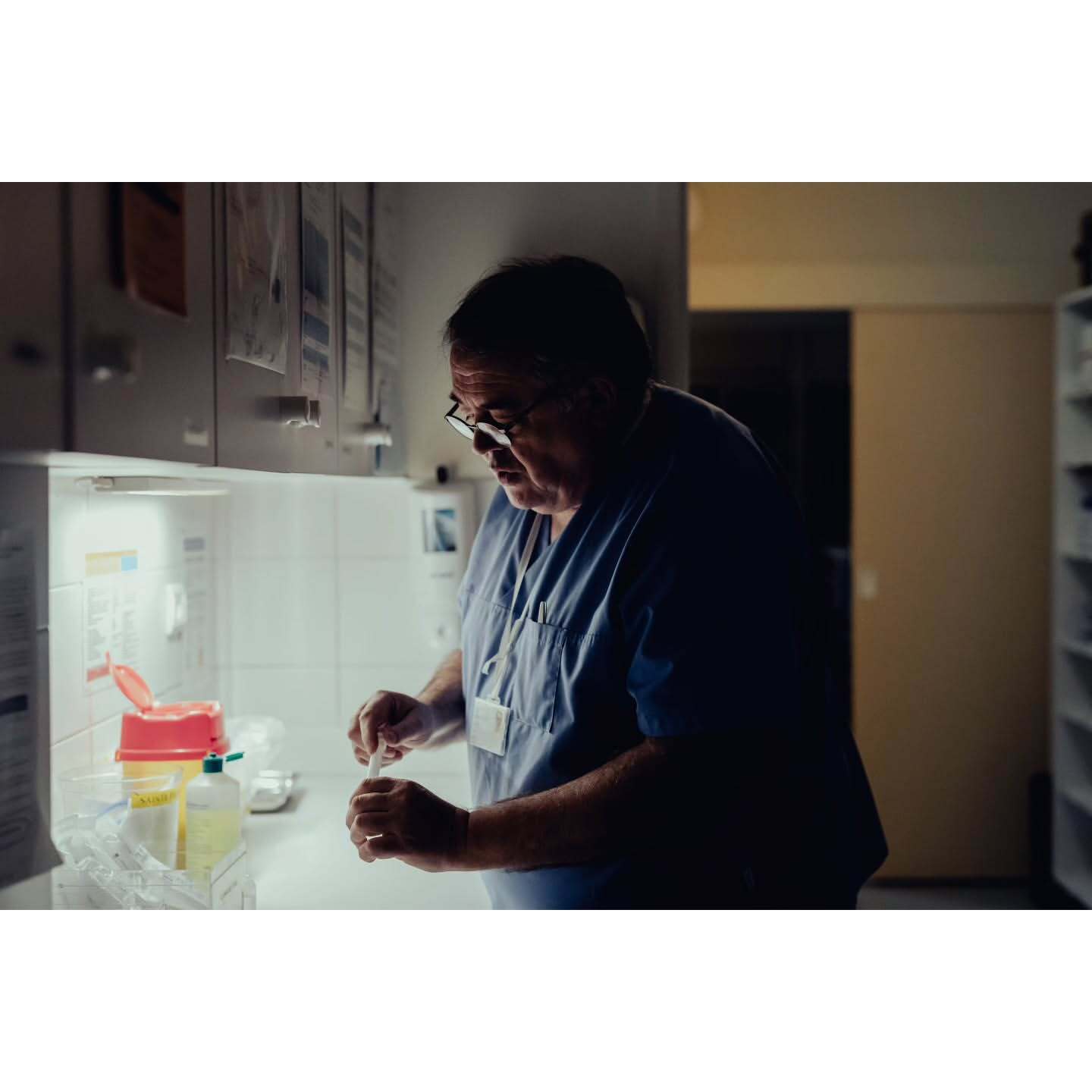 Mature adult male healthcare professional in scrubs prepares medication in a brightly lit indoor clinical environment with medical supplies.