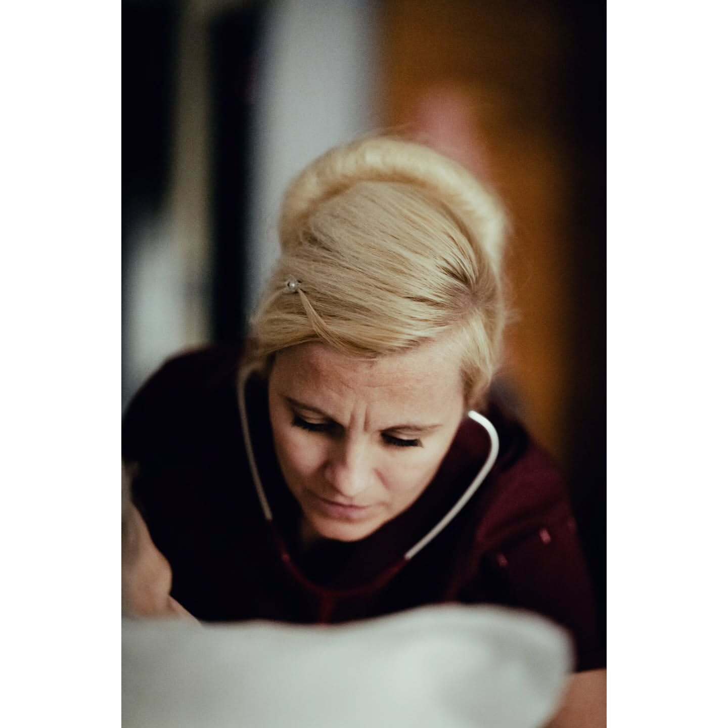 Woman with blonde hair and dark shirt wearing a stethoscope, looking down in an indoor setting with soft lighting.