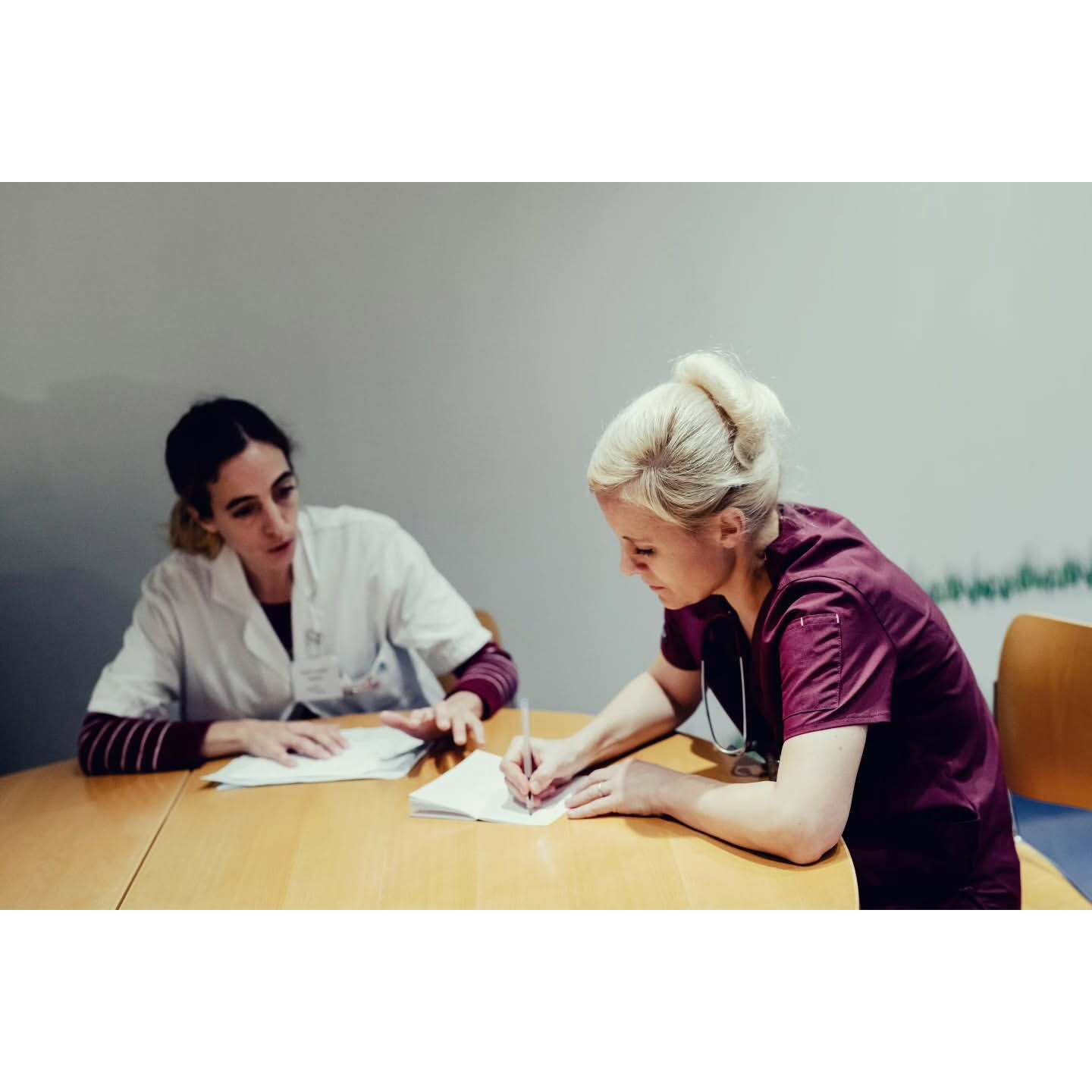 Two female healthcare workers in scrubs and lab coat collaborate at a wooden table, taking notes on papers in a clinical setting.