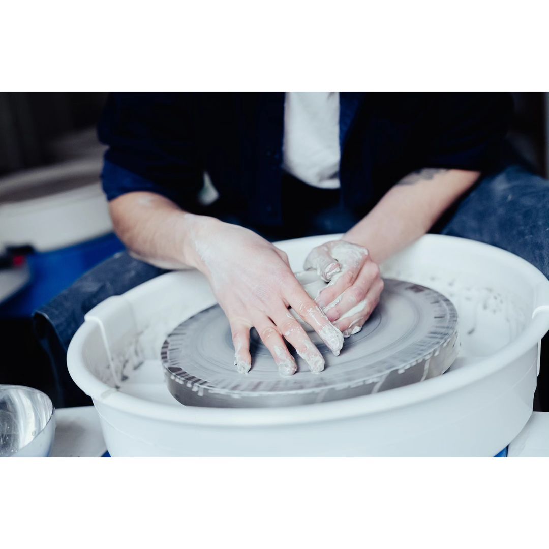 Person's hands shaping clay on a pottery wheel in a bright, indoor studio setting. Focus on the craft and hands-on process.