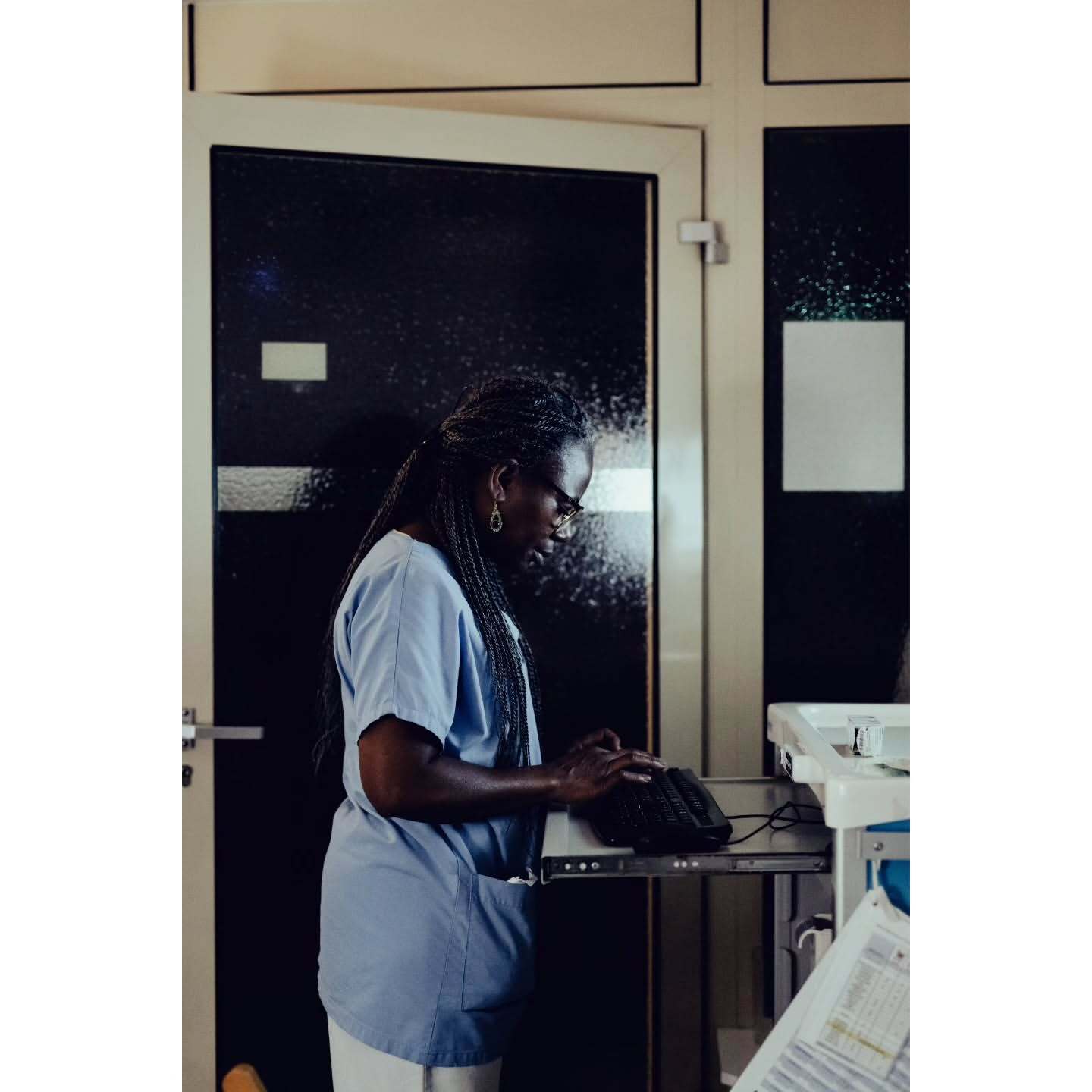 Healthcare worker in scrubs types on a keyboard next to a medical cart in an indoor setting with a dark doorway behind her.