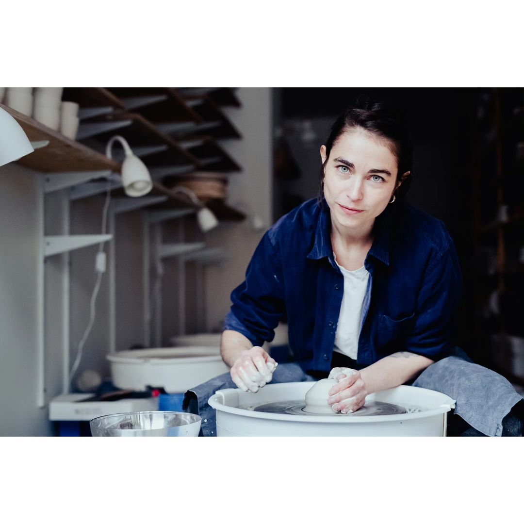 Woman forming clay on a pottery wheel in an indoor studio, wearing a blue shirt, with shelves and pottery in the background.