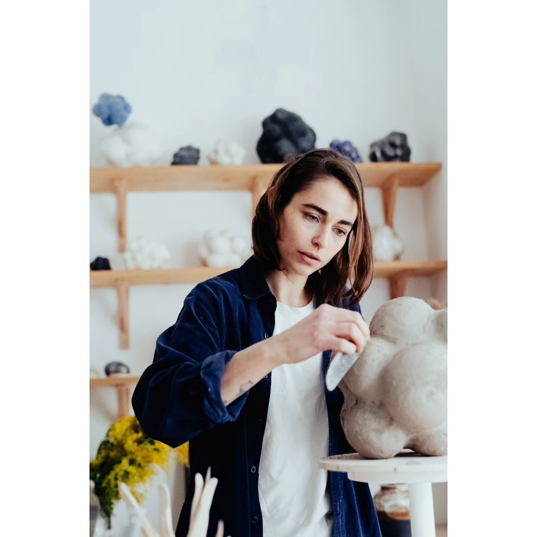 An artist shapes a clay sculpture in a bright studio, using a sculpting tool. The sculpture sits on a stool, surrounded by art supplies.