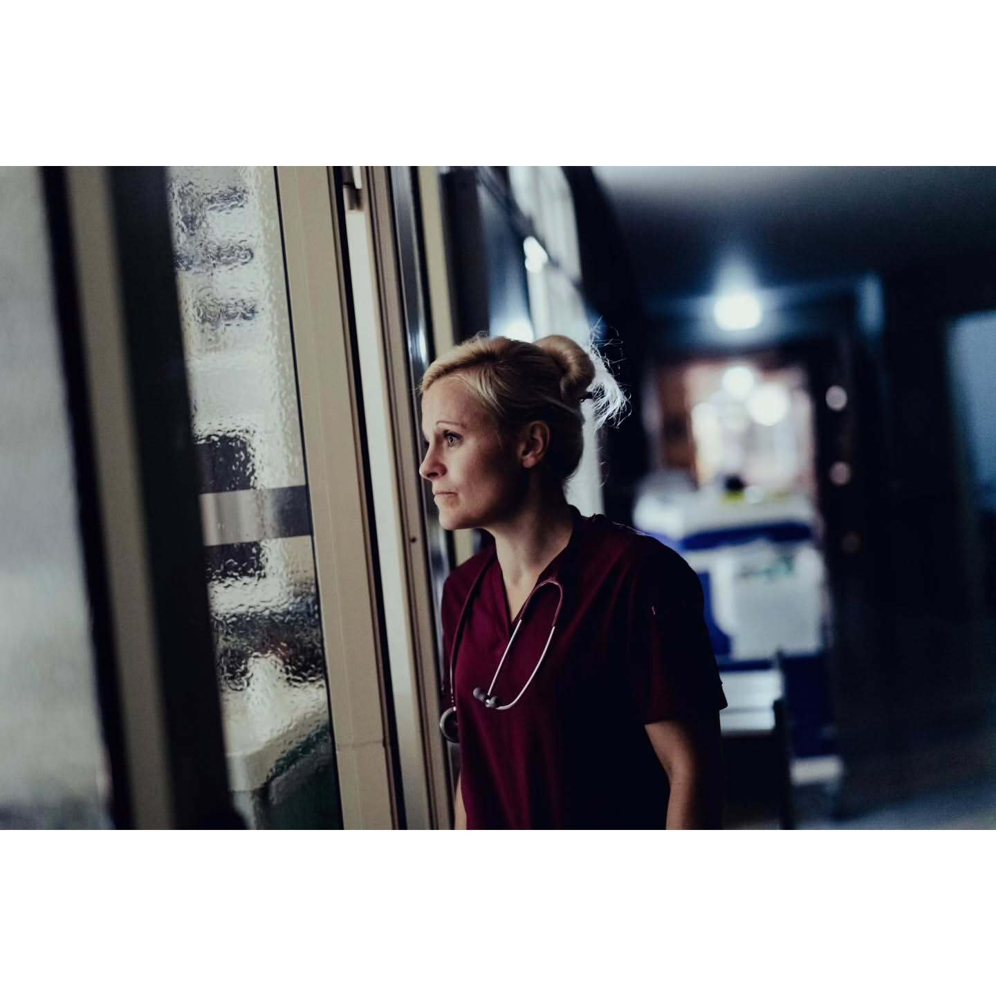 Blonde woman in dark red scrubs and stethoscope looks out of a hospital window at night. She has her hair in a bun, medium shot.