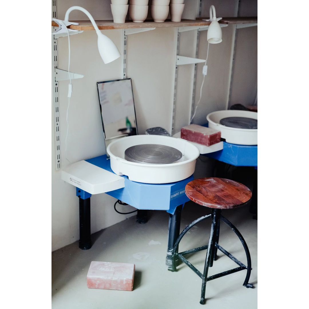 Pottery wheel and stool in an art studio with shelves of clay pots, illuminated by white desk lamps, creating a crafting workspace.