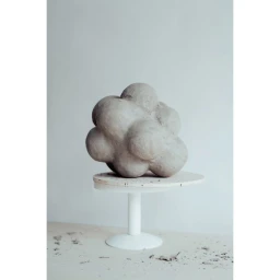 A round, textured grey sculptural art object resting on a white pedestal stand against a plain background in Paris.
