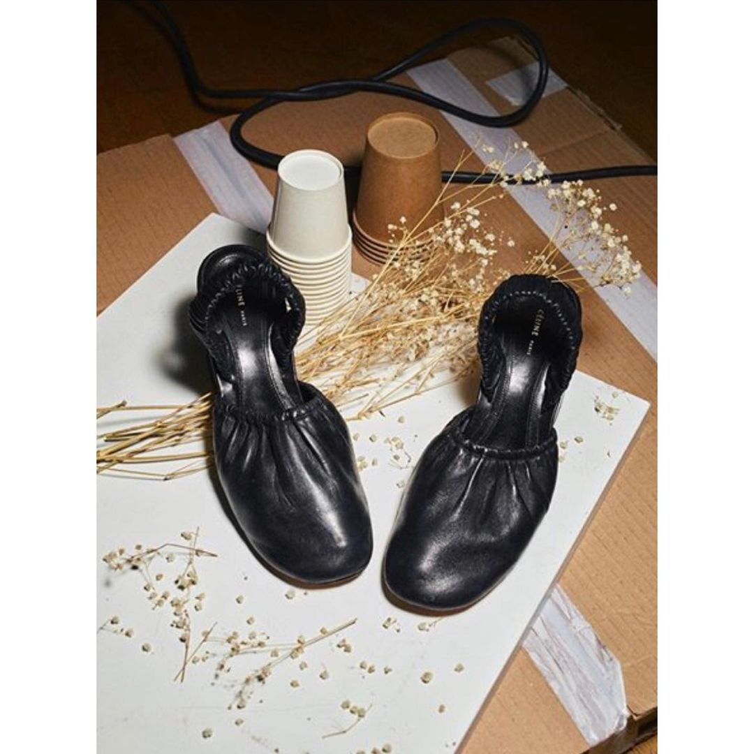 Black heeled mule shoes on a white surface with dried floral accents, craft cups and corrugated cardboard background.
