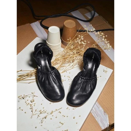 Black heeled mule shoes on a white surface with dried floral accents, craft cups and corrugated cardboard background.