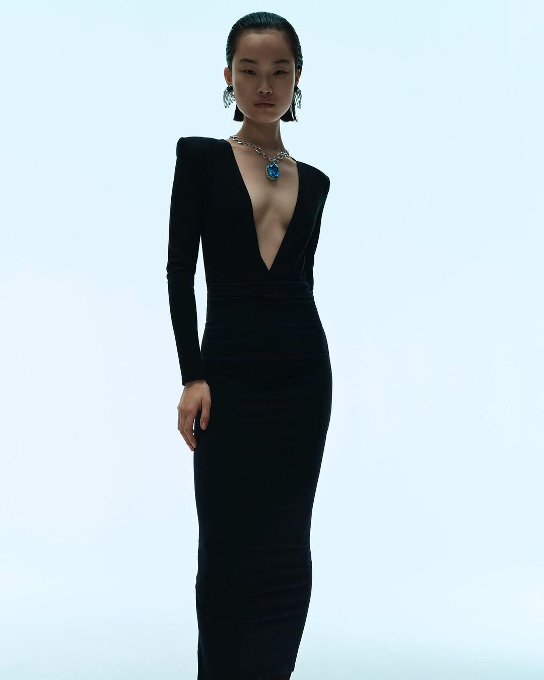 A fashionable Asian woman poses in a long black dress with a plunging neckline and statement jewelry against a bright white backdrop.