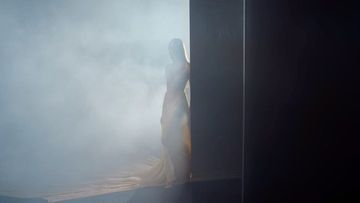 An ethereal woman stands in a cloud of smoke next to a black panel in a dimly lit studio, creating a mysterious and artistic atmosphere.