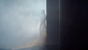An ethereal woman stands in a cloud of smoke next to a black panel in a dimly lit studio, creating a mysterious and artistic atmosphere.