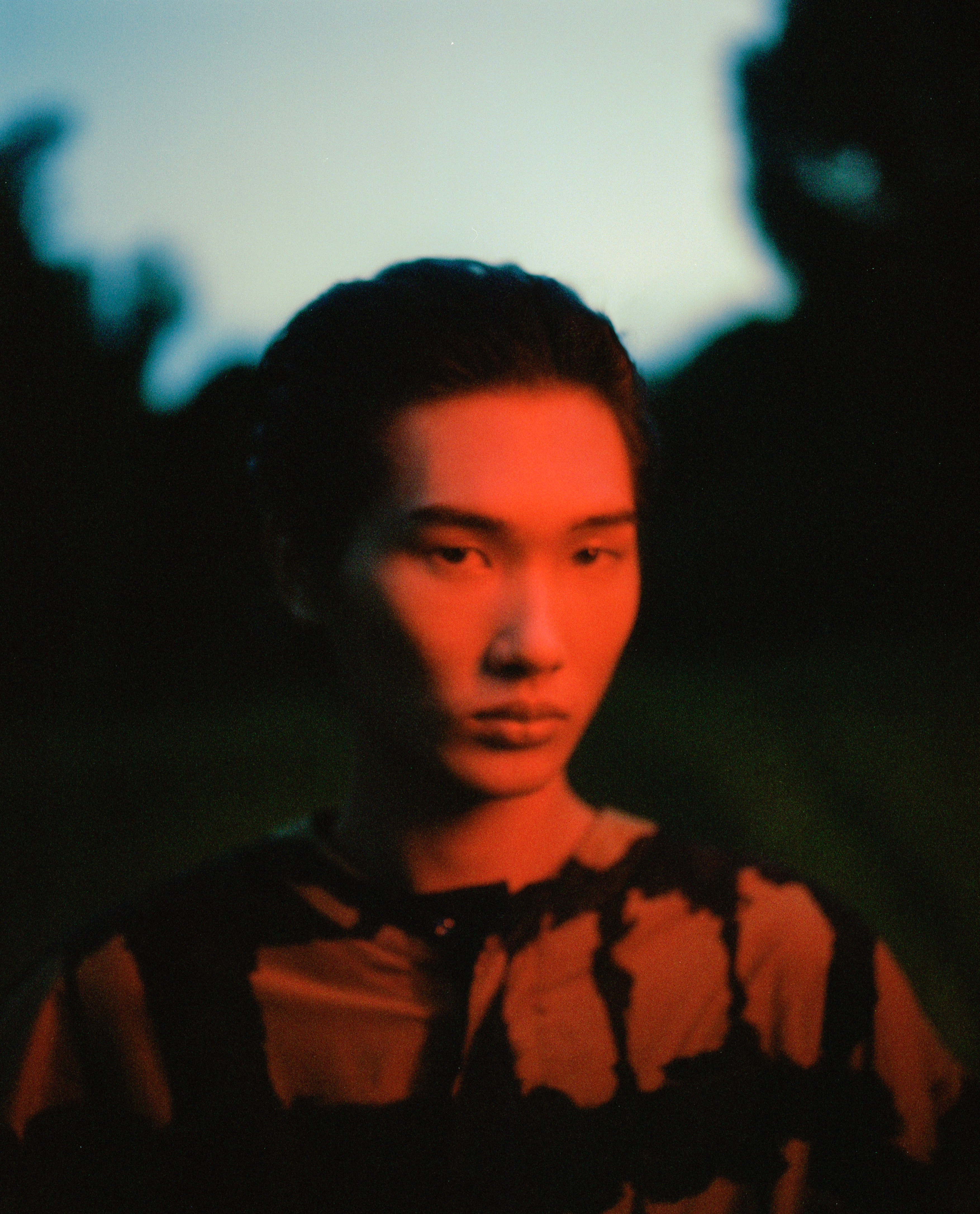 A young East Asian man in a patterned shirt illuminated by vibrant red lighting against a dark twilight outdoor background.