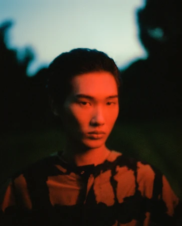 A young East Asian man in a patterned shirt illuminated by vibrant red lighting against a dark twilight outdoor background.