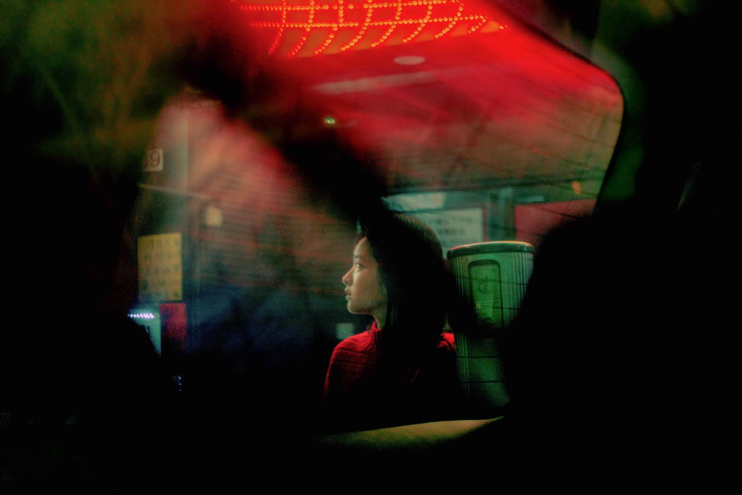 Woman in profile illuminated by red neon reflections looking through a glass window at night.