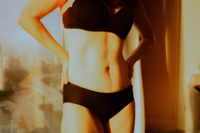 Close up of a woman standing indoors with hands on hips wearing a black bra and matching black underwear in warm sunlight.