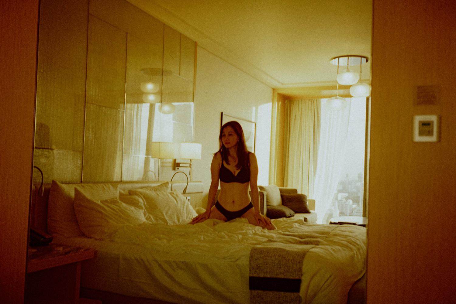 A woman in black lingerie kneeling on an unmade hotel bed illuminated by warm ambient light.