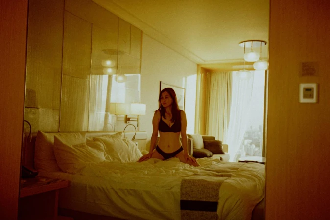 A woman in black lingerie kneeling on an unmade hotel bed illuminated by warm ambient light.