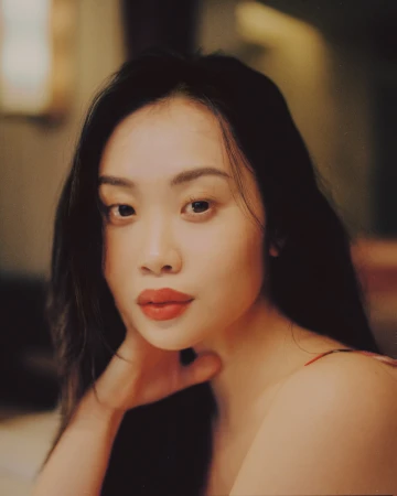 Close-up portrait of an East Asian woman with dark hair and red lipstick resting her hand by her neck against a warm background.