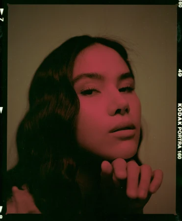Close-up film portrait of a woman with dark wavy hair resting her chin on her hand under red-tinted lighting.