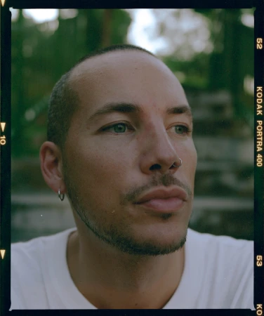 Close-up portrait of a man with green eyes and facial piercings wearing a white t-shirt within a Kodak Portra film border.