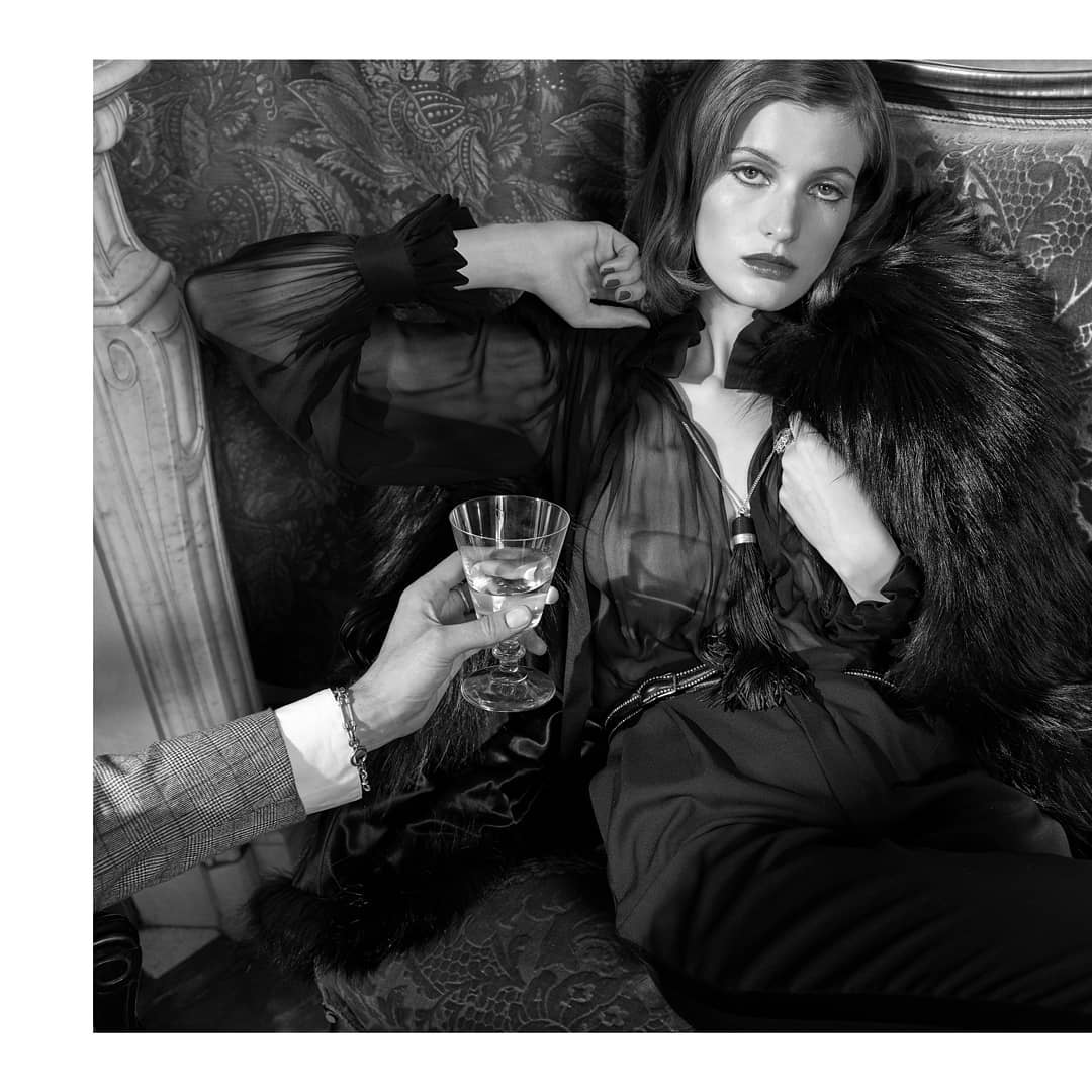 Black and white portrait of a woman in sheer top, holding glass, with arm reaching in, set against patterned background.