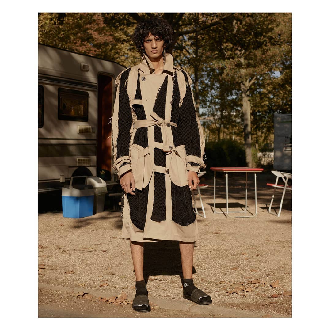 A male model poses in an avant-garde beige and black trench coat with a tie belt in an outdoor campground setting with a trailer.