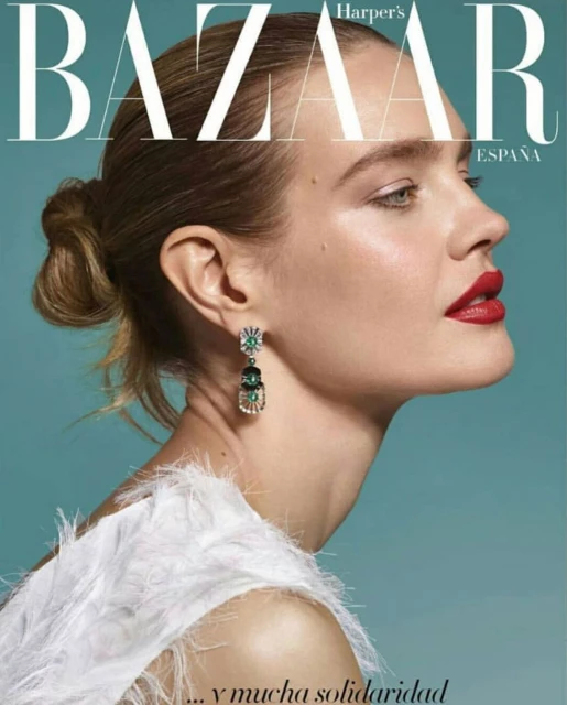 Close-up studio shot of a fair-skinned woman with red lipstick and earrings, wearing feathered attire against a blue background.