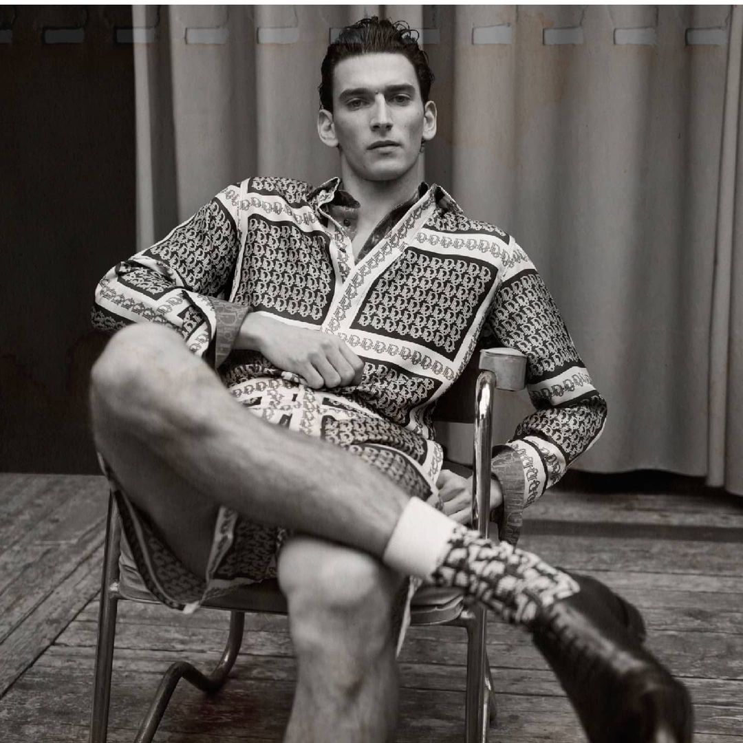 Man in patterned long-sleeve shirt and shorts seated on a metal chair in a monochrome portrait by Nicolas Valois
