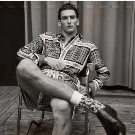 Man in patterned long-sleeve shirt and shorts seated on a metal chair in a monochrome portrait by Nicolas Valois