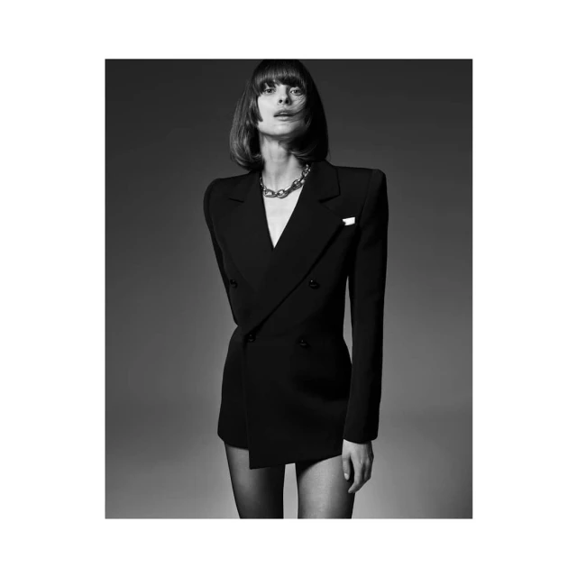 Black and white fashion shot of woman in a double-breasted blazer dress with silver chain necklace, posing against a grey backdrop.