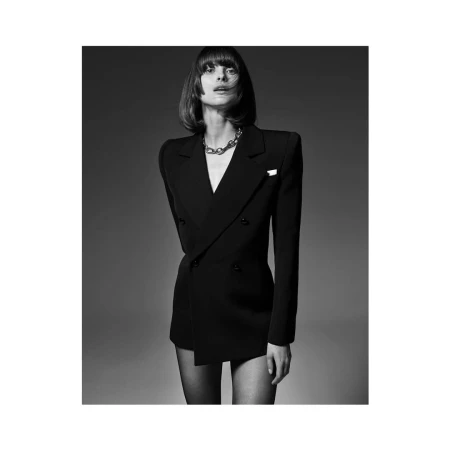 Black and white fashion shot of woman in a double-breasted blazer dress with silver chain necklace, posing against a grey backdrop.