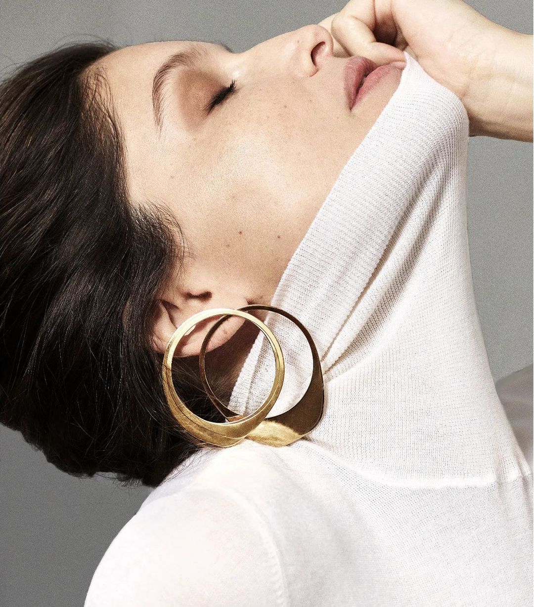 Elegant portrait of a brunette woman with closed eyes wearing a white turtleneck and sculptural gold earrings in a minimalist grey setting.