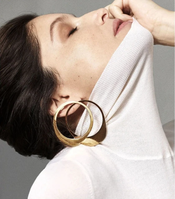 Elegant portrait of a brunette woman with closed eyes wearing a white turtleneck and sculptural gold earrings in a minimalist grey setting.