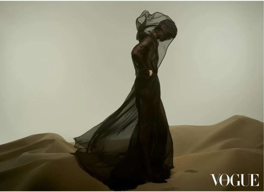 A person draped in black fabric strikes a pose on a sand dune, against a light background in a fashion editorial style image for Vogue.