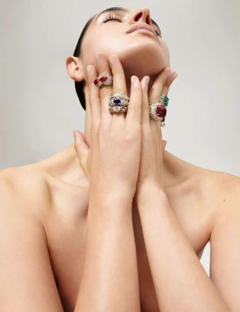Adult female poses with hands raised to her neck, displaying multiple gemstone rings in a beauty shot with diffused, neutral lighting.