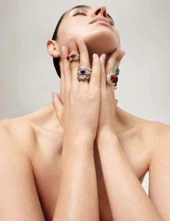 Adult female poses with hands raised to her neck, displaying multiple gemstone rings in a beauty shot with diffused, neutral lighting.