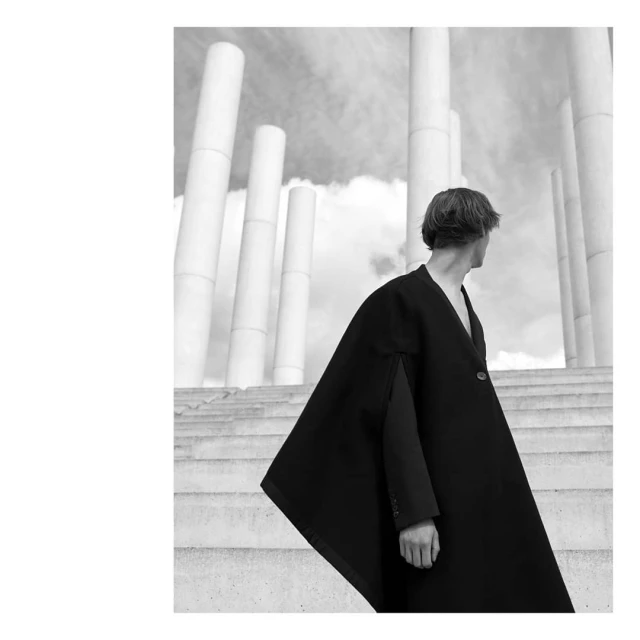 Monochrome fashion shot of a person wearing a dark cloak and jacket on concrete steps with tall, cylindrical columns in the background.