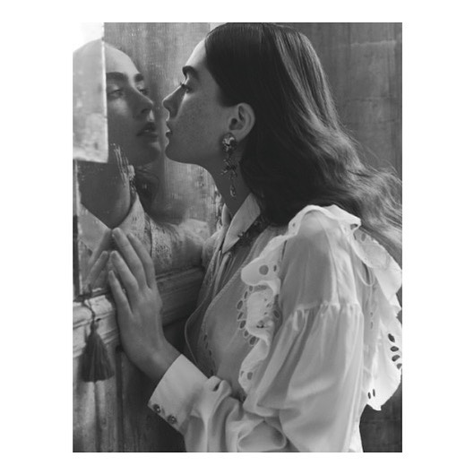 Monochrome portrait of a woman standing close to a mirror, her face nearly touching the glass, wearing an eyelet blouse and earrings.