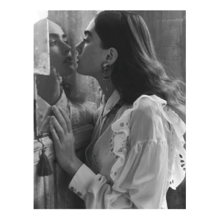 Monochrome portrait of a woman standing close to a mirror, her face nearly touching the glass, wearing an eyelet blouse and earrings.