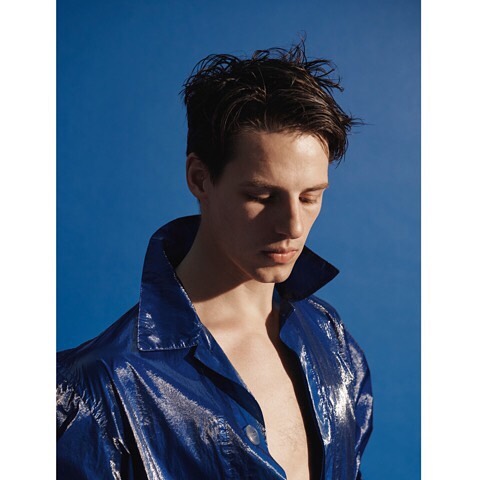 Portrait of a young adult man in a blue, shiny jacket against a bright blue backdrop, captured in a medium shot with soft lighting.