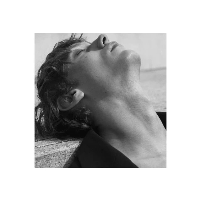 Monochrome close-up of a young adult male with tousled hair, lying with his head tilted back, face toward the sky, wearing a dark jacket.