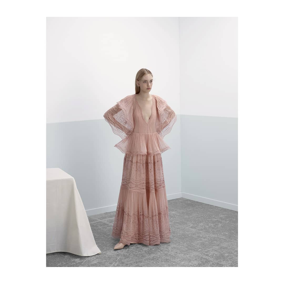 A fair-skinned woman poses in a pale pink gown with tiers and ruffles, accented by a neutral background, in a studio-lit fashion shot.