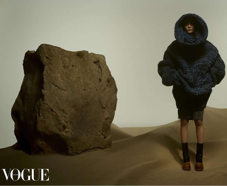 Fashion editorial featuring a person in oversized knitwear next to a large rock formation in a desert-like setting for Vogue magazine.