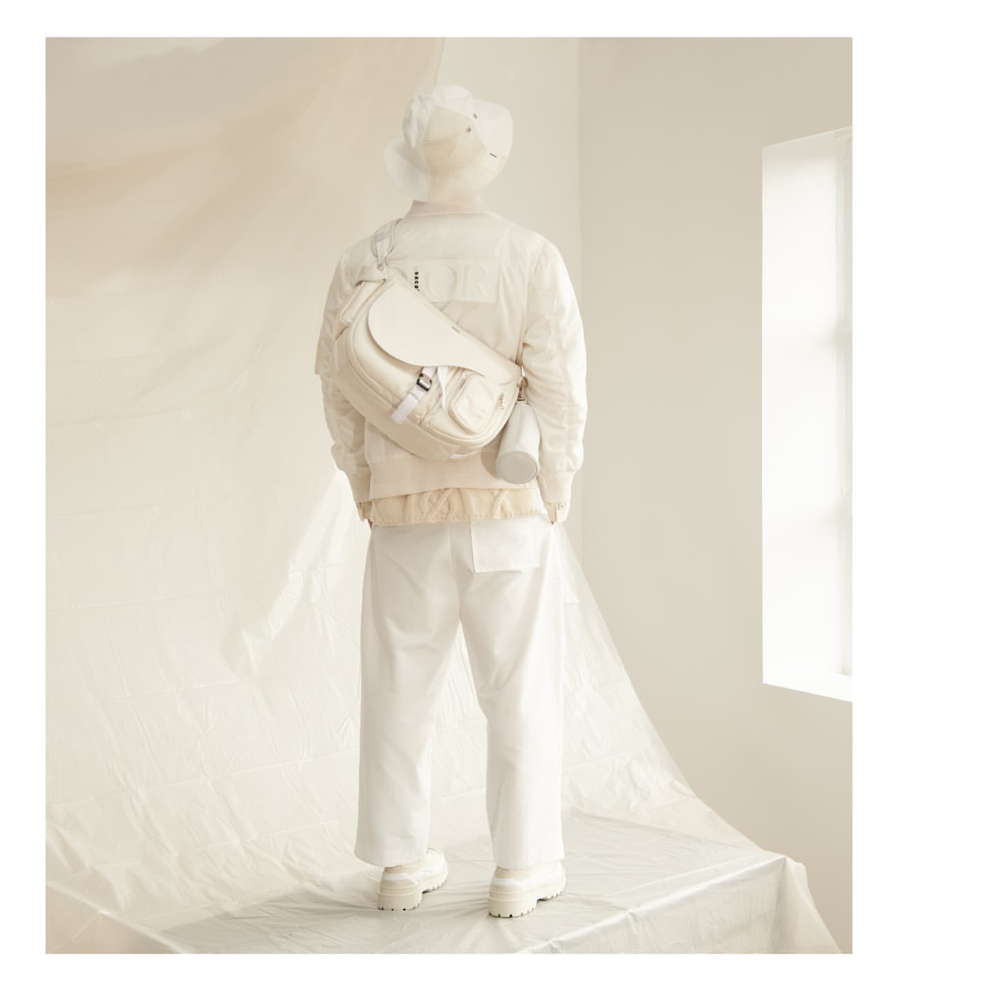 A person from behind in all white clothing, with a white bag and cylinder slung over their shoulder, stands in front of a window.