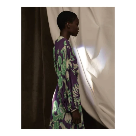 Profile view of a Black woman with short hair wearing a purple dress with green floral pattern in front of a draped fabric backdrop.